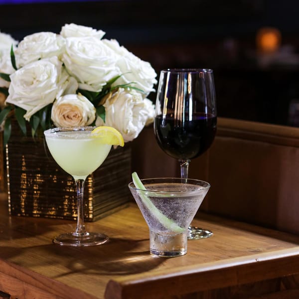 Three drinks, including a margarita and red wine, on a wooden table with white roses.