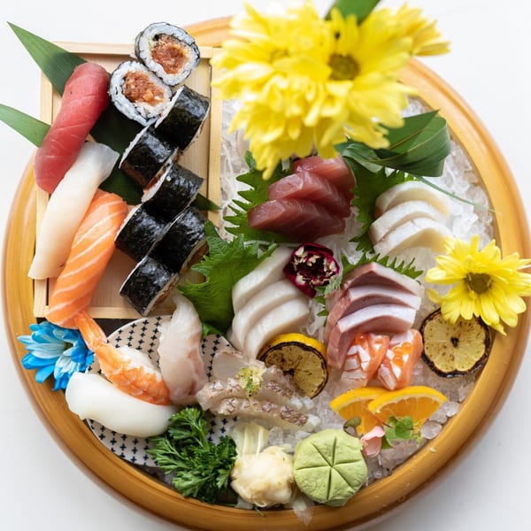 A large wooden platter filled with various sushi, sashimi, and rolls, garnished with flowers and citrus.