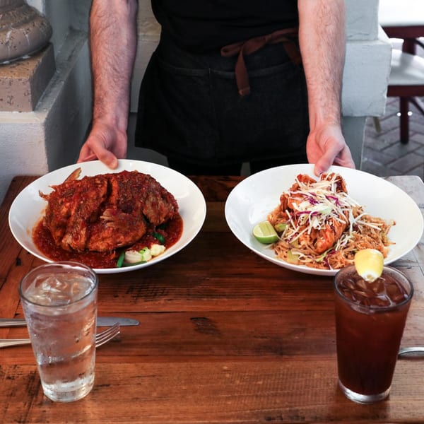 A server presents two Asian dishes, a whole fish in red sauce and pad Thai, with drinks.