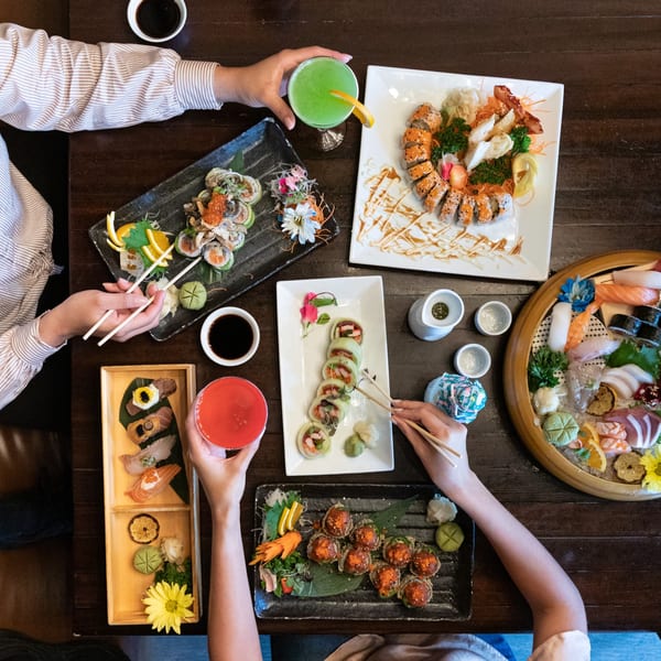 Overhead view of a table laden with various sushi and sashimi platters, cocktails, and diners' hands.