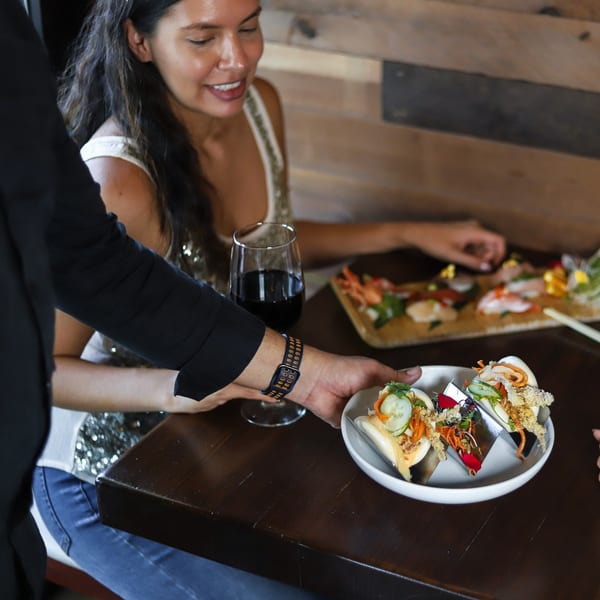 A server presents a plate of bao buns to a smiling woman at a table with sushi and wine.