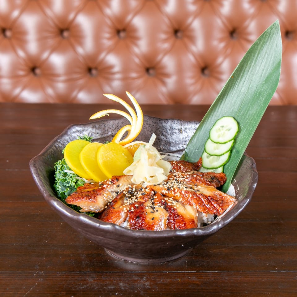 Unagi donburi with grilled eel, pickled radish, ginger, and cucumber, on a wooden table.