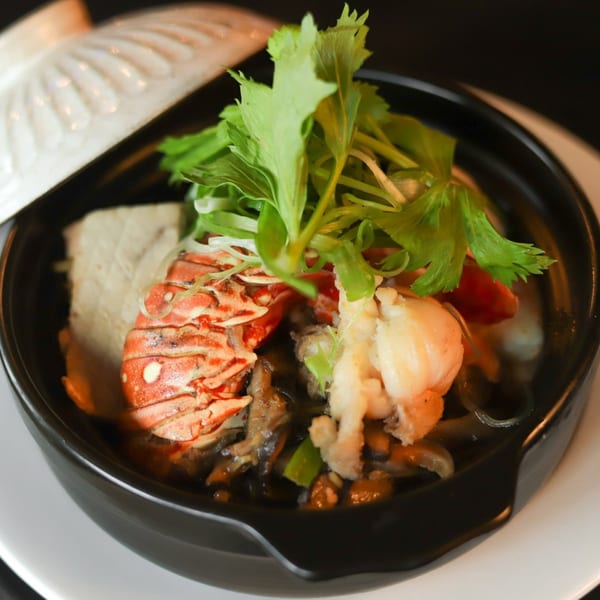 A black bowl of Asian cuisine with lobster, shrimp, and green herbs, served with a white lid.